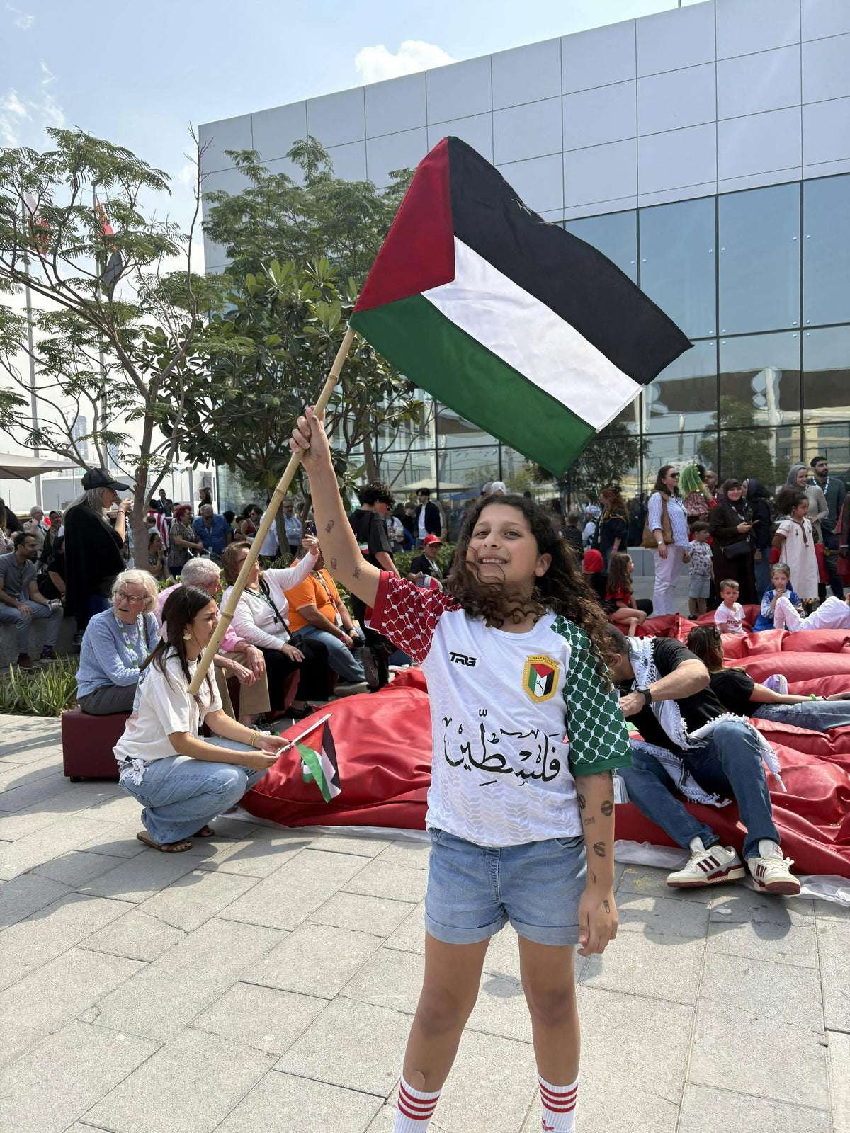 Girl wearing Palestine football jersey white short sleeve waving Palestine flag at solidarity rally