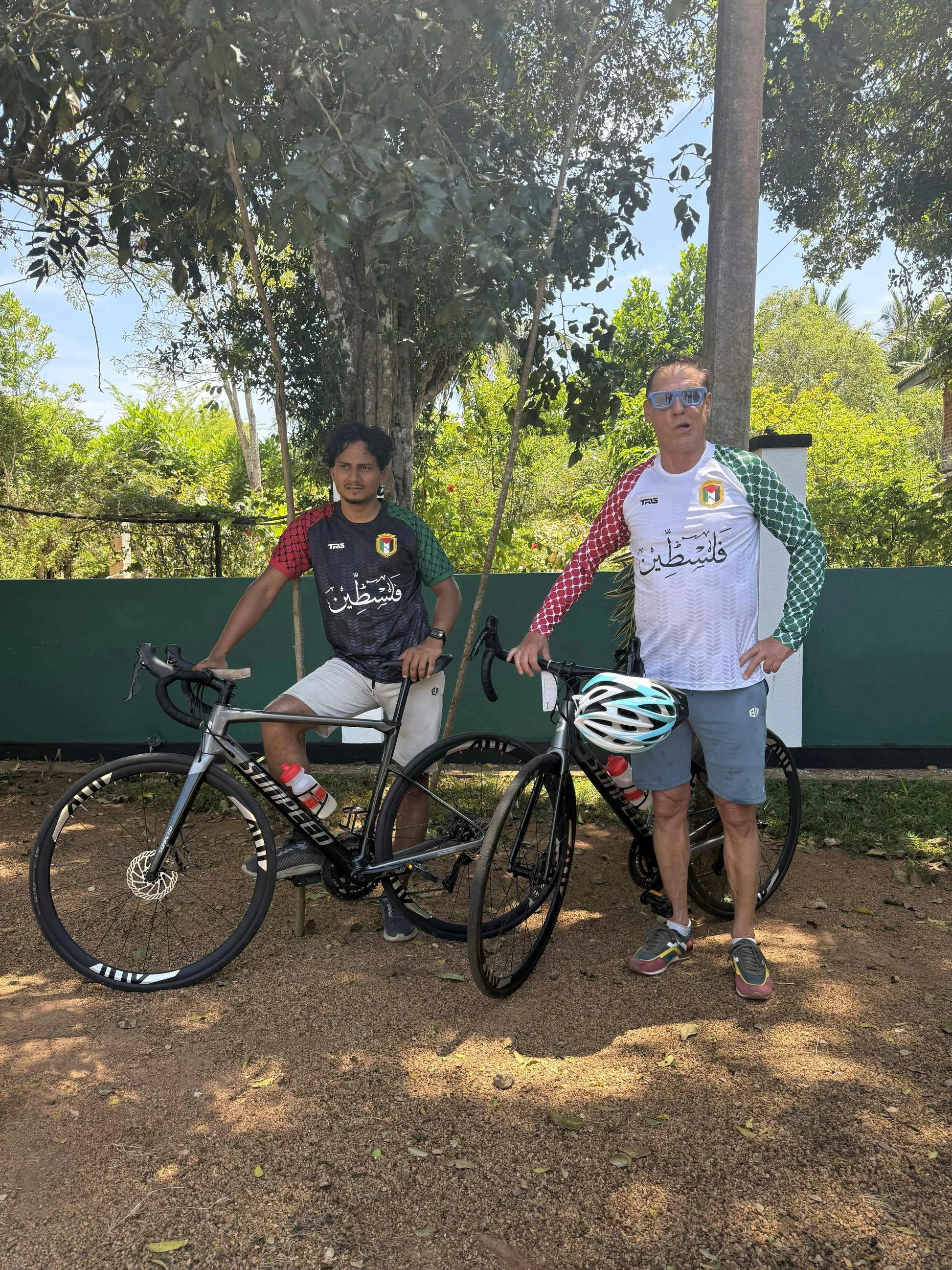 Two men wearing Palestine football jerseys black and white long sleeve with bicycles outdoors in Sri Lanka